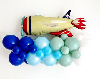 Könnte beinhalten: Ein roter, weißer und blauer Flugzeugballon mit einem Flammenschwanz, umgeben von einer Girlande aus blauen, hellblauen und grünen Ballons.