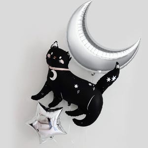 May include: A black cat-shaped foil balloon with white star patterns and a pink collar featuring a crescent moon charm, accompanied by a large silver crescent moon balloon and a smaller silver star-shaped balloon, all inflated and floating.