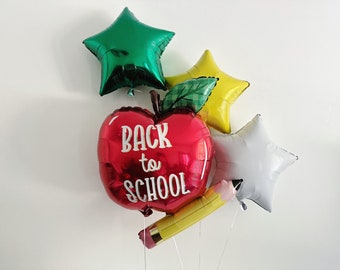 May include: A collection of foil balloons, including a red apple with "BACK to SCHOOL" text, a yellow pencil, and three star-shaped balloons in green, yellow, and white. The balloons are tied together with white string.