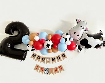 May include: A black number two balloon, a cow balloon, and a garland of balloons in red, blue, brown, and white with black and white cow print. The garland has a banner that says "Moo Moo I'm Two".