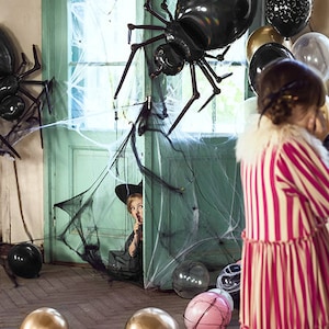May include: A black spider balloon with long legs hangs in a doorway decorated with spiderwebs. A child wearing a witch hat peeks out from behind the door. There are other balloons in the background, including gold, silver, and black.