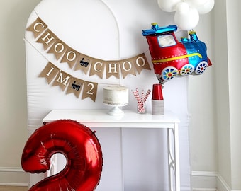 May include: A red number two balloon, a white table with a cake and red and white striped cups, a banner that says "Choo Choo I'm 2" and a red, blue, and yellow train balloon with white clouds above it.