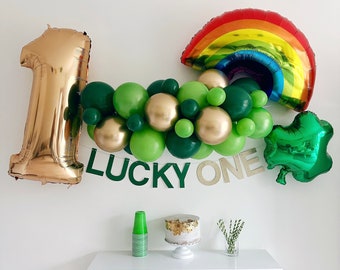 May include: Gold number one balloon, green and gold balloons, a rainbow balloon, and a green shamrock balloon with the text "LUCKY ONE" in green letters.