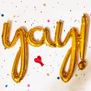 Könnte beinhalten: Goldene Folien-Buchstabenballons, die "yay!" buchstabieren, mit einem roten Herzen auf weißem Hintergrund mit Konfetti.