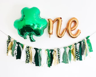 May include: Green shamrock foil balloon and gold foil "one" balloon with a green, white, and gold fabric banner with shamrock and polka dot patterns.