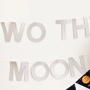 K&ouml;nnte beinhalten: Ein silbernes Glitzerbanner mit der Aufschrift "TWO THE MOON" an einer wei&szlig;en Wand. Darunter ein kleines schwarzes Wimpel mit einer Planeten-Grafik und dem Wort "Jupiter". Auf der rechten Seite ist ein silberner Ballon zu sehen.
