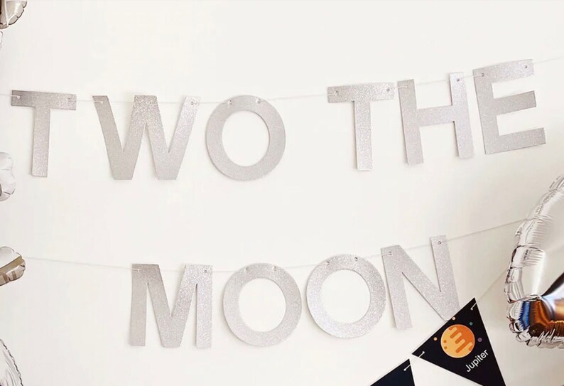 K&ouml;nnte beinhalten: Ein silbernes Glitzerbanner mit der Aufschrift "TWO THE MOON" an einer wei&szlig;en Wand. Darunter ein kleines schwarzes Wimpel mit einer Planeten-Grafik und dem Wort "Jupiter". Auf der rechten Seite ist ein silberner Ballon zu sehen.