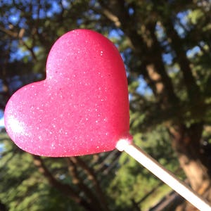 May include: A pink heart-shaped lollipop on a white stick. The lollipop is covered in glitter and is in focus, while the background is blurred.