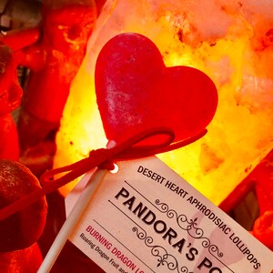 May include: A red heart-shaped lollipop on a stick with a white label that reads "DESERT HEART APHRODISIAC LOLLIPOPS PANDORA'S POPS BURNING DRAGON LOVE Roaring Dragon Fruit and Tender Cherry NET WT 0.6 OZ (18 g)"
