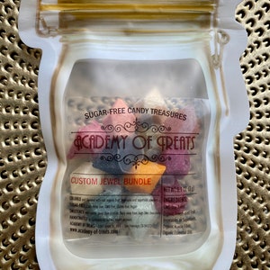 May include: A clear plastic bag with a zipper closure, containing a variety of colorful sugar-free candy treats. The bag is shaped like a jar with a gold lid. The candy is in the shape of stars and is labeled "SUGAR-FREE CANDY TREASURES" and "ACADEMY OF GREATS CUSTOM JEWEL BUNDLE". The bag also includes the ingredients and net weight of the candy.