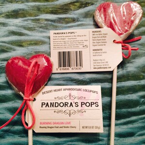 May include: Two heart-shaped lollipops on sticks. The lollipops are red and have a clear plastic coating. The lollipops are tied with a pink ribbon. The lollipop on the left has a label that says "Burning Dragon Love" and "Roaring Dragon Fruit and Tender Cherry". The lollipop on the right has a label that says "Pandora's Pops" and lists the ingredients. The label also includes a barcode and the company's website.
