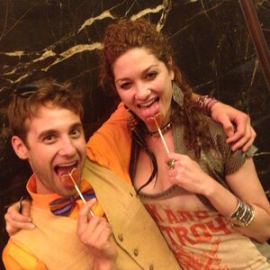 May include: Two people are holding and eating heart-shaped lollipops. The person on the left is wearing a tan vest, a blue and orange bow tie, and an orange shirt. The person on the right is wearing a grey shirt with the text "Ramp Troy" printed on it.