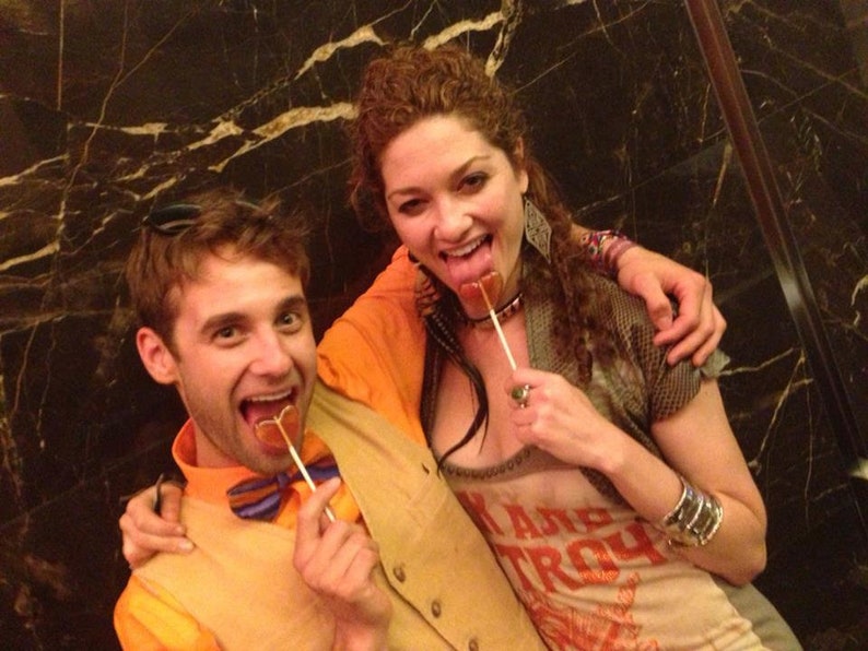 May include: Two people are holding and eating heart-shaped lollipops. The person on the left is wearing a tan vest, a blue and orange bow tie, and an orange shirt. The person on the right is wearing a grey shirt with the text "Ramp Troy" printed on it.