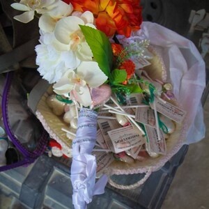 May include: A white and orange bridal bouquet with white orchids, orange flowers, and green leaves. The bouquet is wrapped in white satin ribbon and is sitting on a basket of white and pink candy.