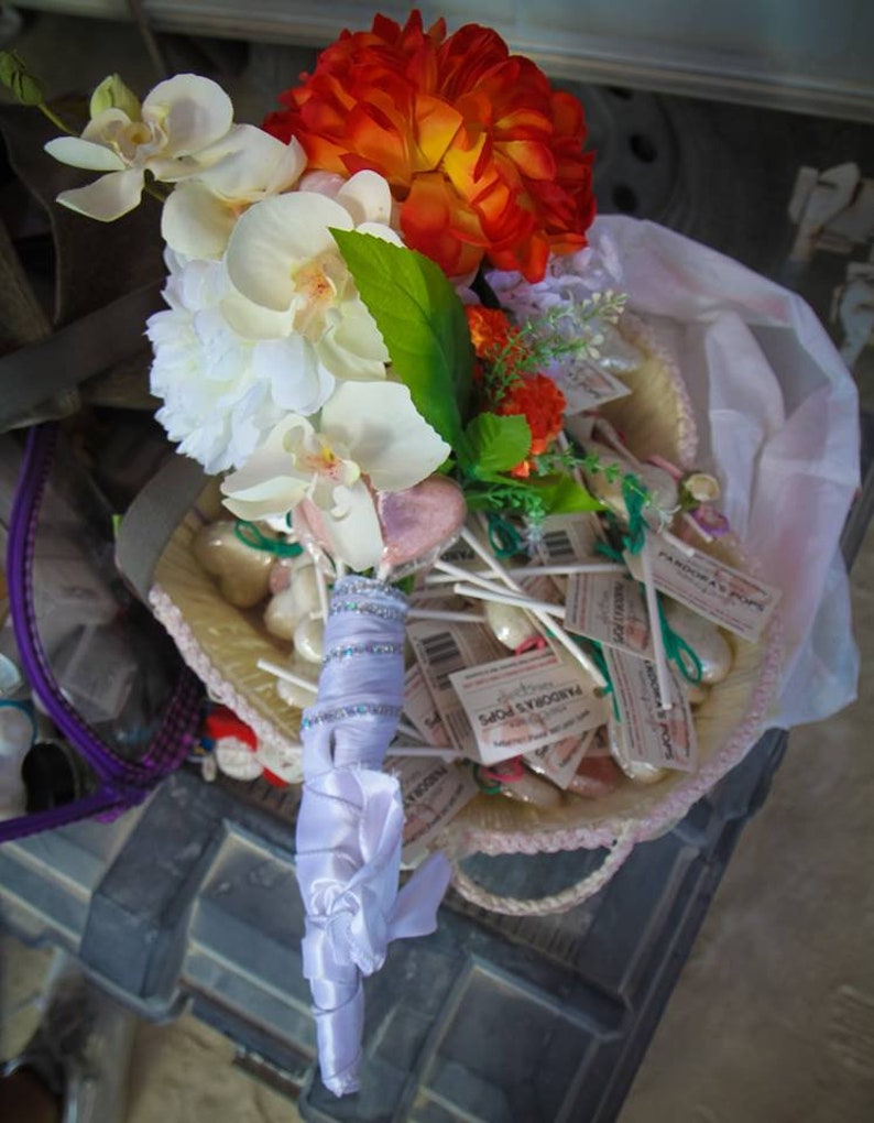 May include: A white and orange bridal bouquet with white orchids, orange flowers, and green leaves. The bouquet is wrapped in white satin ribbon and is sitting on a basket of white and pink candy.