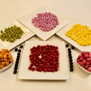 May include: Six small white bowls filled with colorful candy shaped like diamonds. The candy is in shades of pink, red, yellow, green, and orange. There are also black, diamond-shaped candies in a row on the white surface.