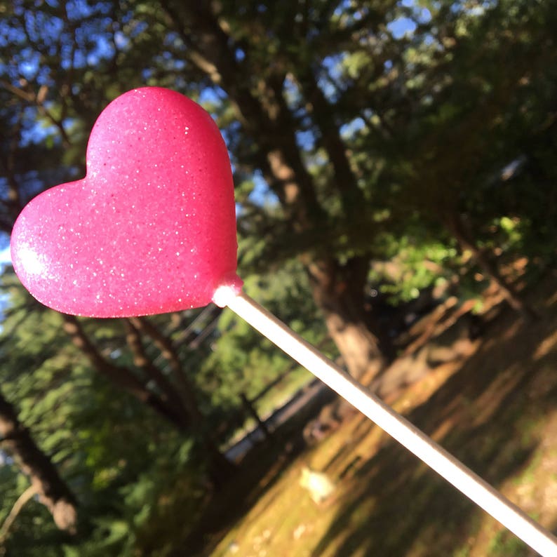 May include: A pink heart-shaped lollipop on a white stick. The lollipop is covered in glitter and is in focus, while the background is blurred.
