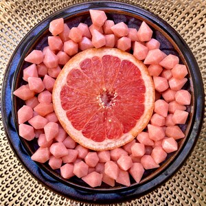 May include: A plate of pink candy shaped like diamonds surrounds a slice of grapefruit. The plate is on a gold background.