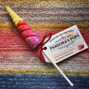 May include: A pink, red, and yellow candy unicorn horn on a stick with a white tag that reads "BURNING MAN LOVE CANDY PLAYTOY PANDORA'S POPS TRANSFORMATION TICKLER Opening Minds & Twisting Flavors".