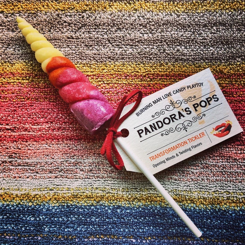 May include: A pink, red, and yellow candy unicorn horn on a stick with a white tag that reads "BURNING MAN LOVE CANDY PLAYTOY PANDORA'S POPS TRANSFORMATION TICKLER Opening Minds & Twisting Flavors".
