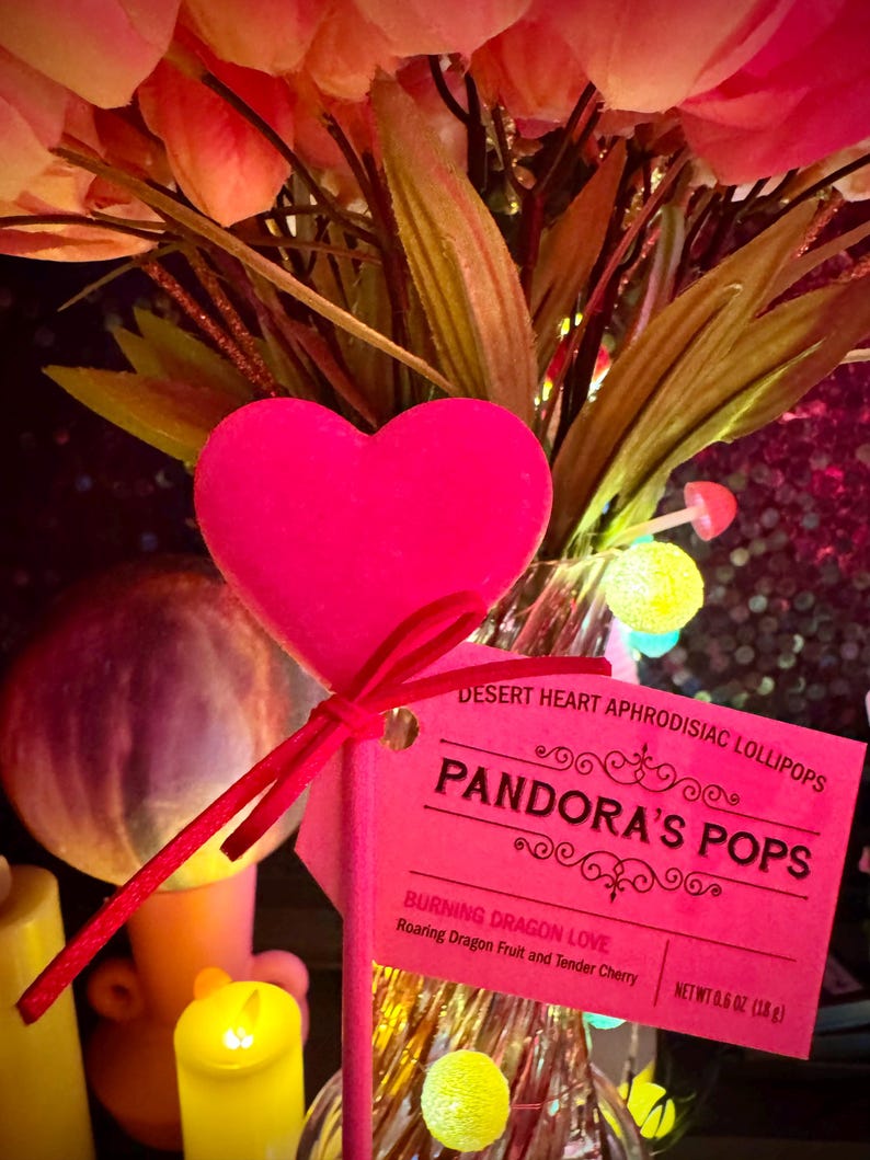 May include: Pink heart-shaped lollipop on a stick with a pink tag that reads "DESERT HEART APHRODISIAC LOLLIPOPS PANDORA'S POPS BURNING DRAGON LOVE Roaring Dragon Fruit and Tender Cherry NET WT 0.6 OZ (18 g)".