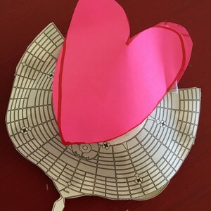 May include: A pink paper heart shape on top of a white paper grid with the words "Radial Road" and "8.00" printed on it.