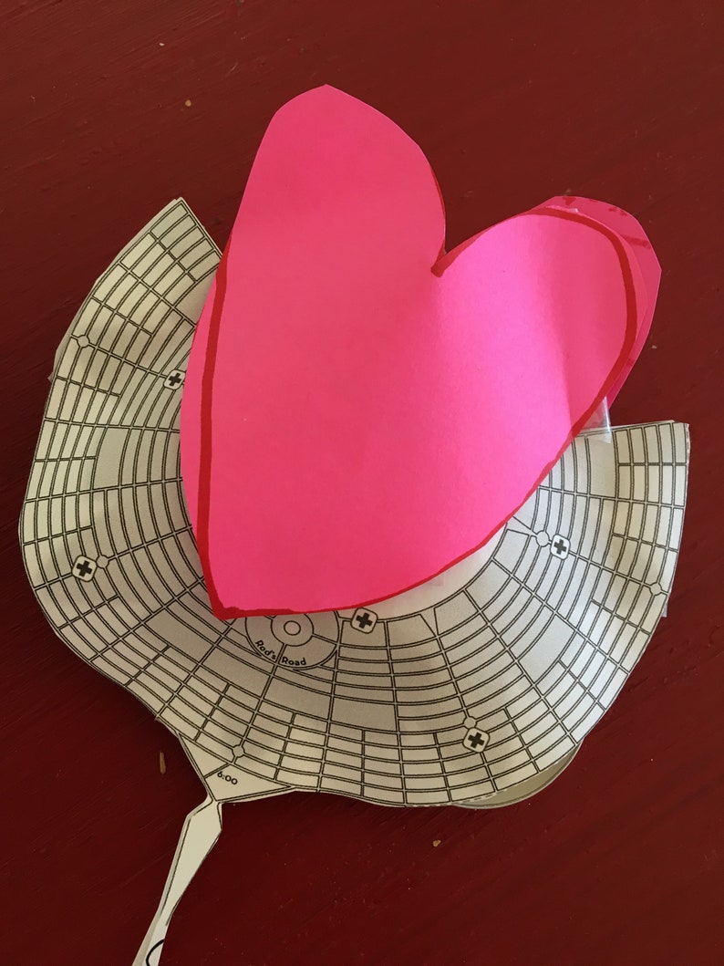 May include: A pink paper heart shape on top of a white paper grid with the words "Radial Road" and "8.00" printed on it.