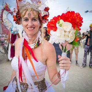 May include: A bride wearing a white dress with a floral crown and a bouquet of red and white flowers. She is standing in a desert setting with other people in the background.