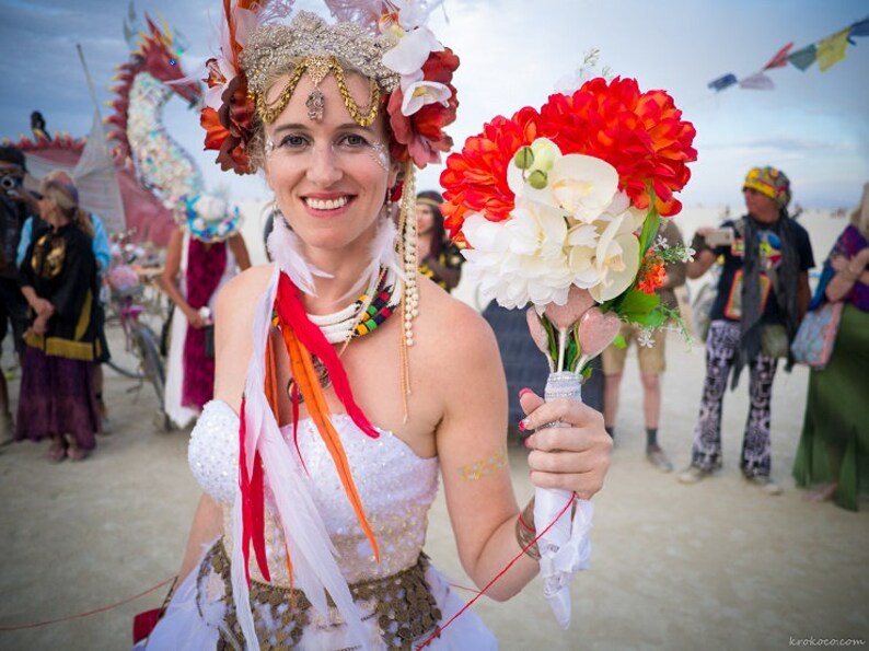 May include: A bride wearing a white dress with a floral crown and a bouquet of red and white flowers. She is standing in a desert setting with other people in the background.