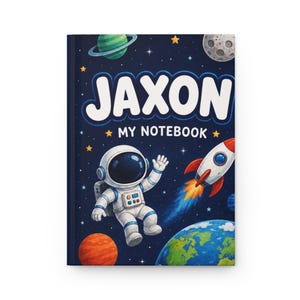 May include: A hardcover notebook with a dark blue space-themed cover featuring the name JAXON and the text MY NOTEBOOK in white. The design includes a white astronaut, a red and white rocket, a green planet with rings, a moon, and Earth.
