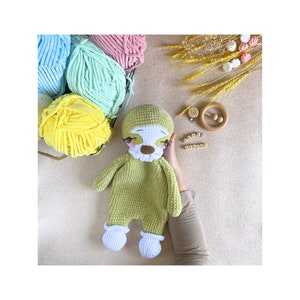 May include: A green crocheted sloth toy with white feet and a white face. The sloth has black eyes and a pink nose. The toy is lying on a beige surface.