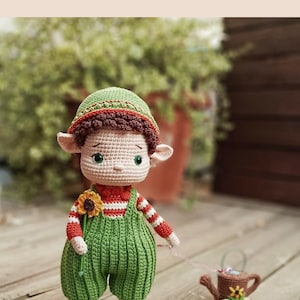 Puede incluir: Muñeca de ganchillo hecha a mano con sombrero verde, peto y zapatos. La muñeca tiene una camisa a rayas rojas y blancas y un girasol. Tira de una pequeña regadera marrón sobre ruedas.