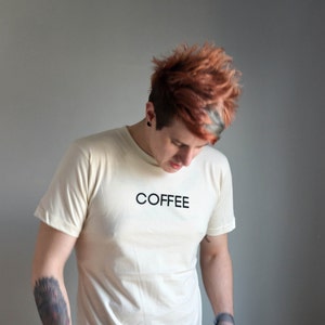 Light coloured, short sleeve t-shirt screen printed to say Coffee in dark font, modelled with dark pants.