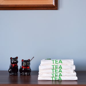 Light coloured, short sleeve t-shirts with bright coloured ringer trim at neck and cuff screen printed to say Tea in matching colour print, stacked neatly next to kitschy milk and sugar serving dishes.