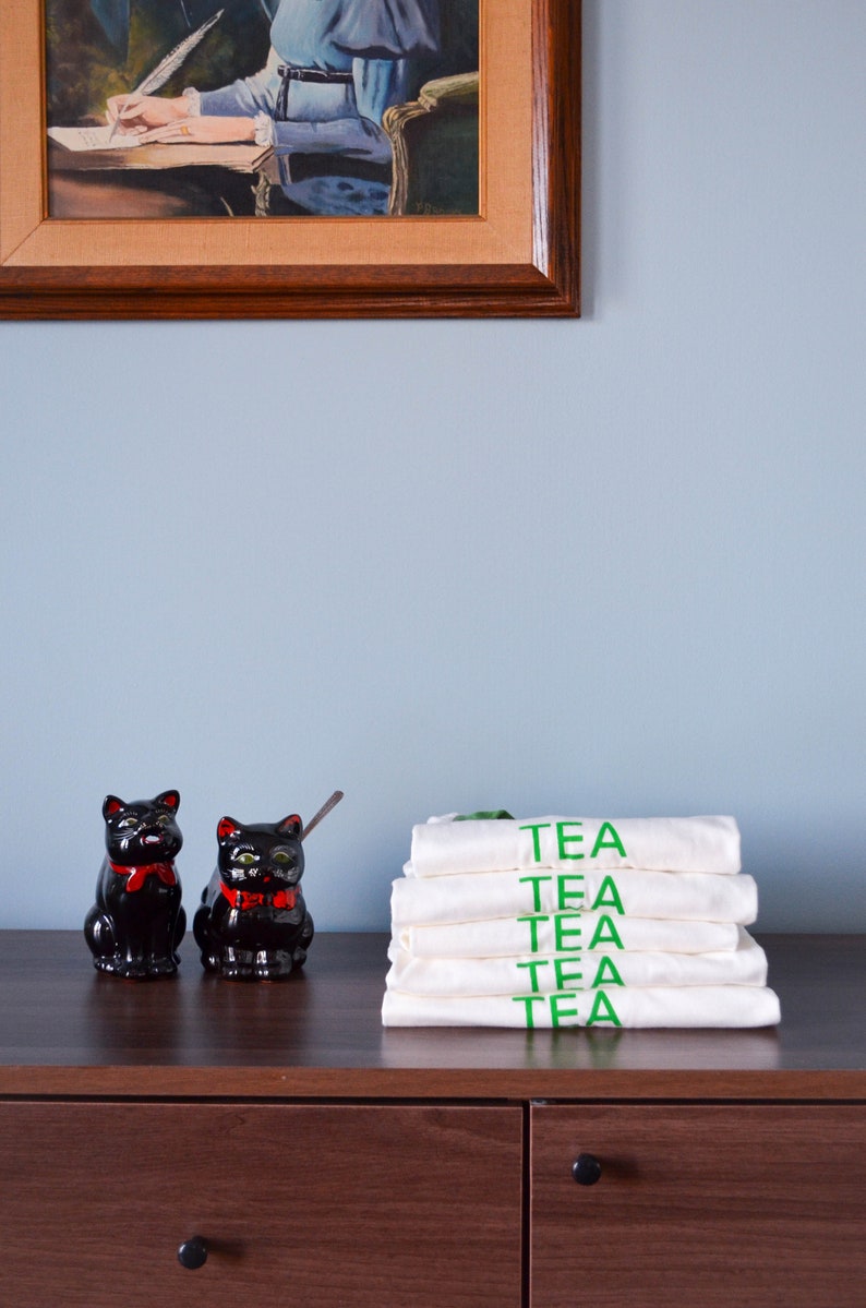 Light coloured, short sleeve t-shirts with bright coloured ringer trim at neck and cuff screen printed to say Tea in matching colour print, stacked neatly next to kitschy milk and sugar serving dishes.