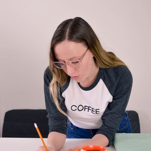 Raglan, three-quarter sleeve t-shirt in contrasting dark sleeve and light body, screen printed to say Coffee in dark ink, modelled by woman in jeans.