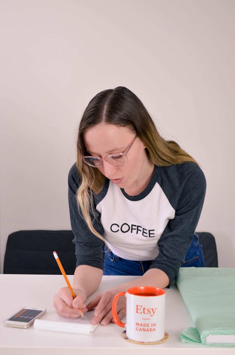 Raglan, three-quarter sleeve t-shirt in contrasting dark sleeve and light body, screen printed to say Coffee in dark ink, modelled by woman in jeans.