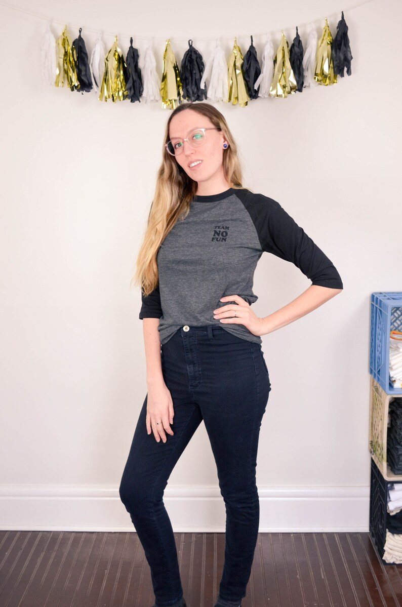 Raglan half length sleeve t-shirt in dark colours, screen printed to say Team No Fun in small type, modelled in dark pants and shoes to show it tucked in.