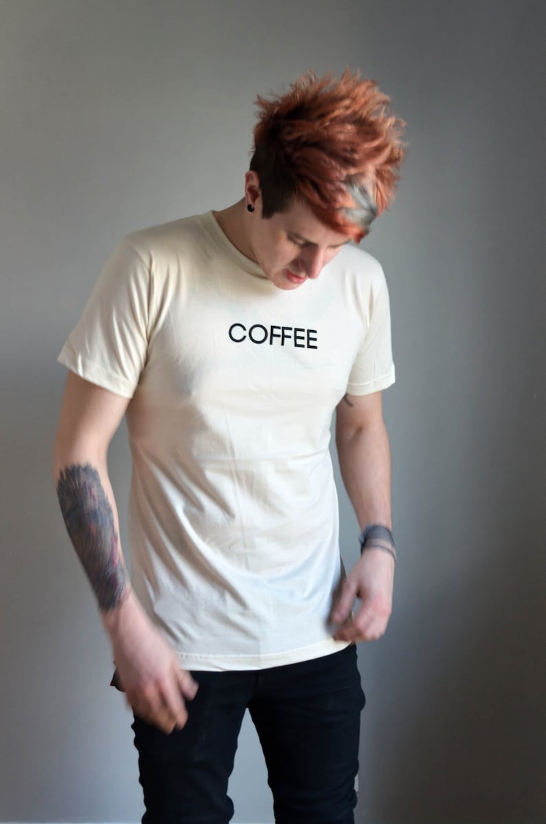 Light coloured, short sleeve t-shirt screen printed to say Coffee in dark font, modelled with dark pants.