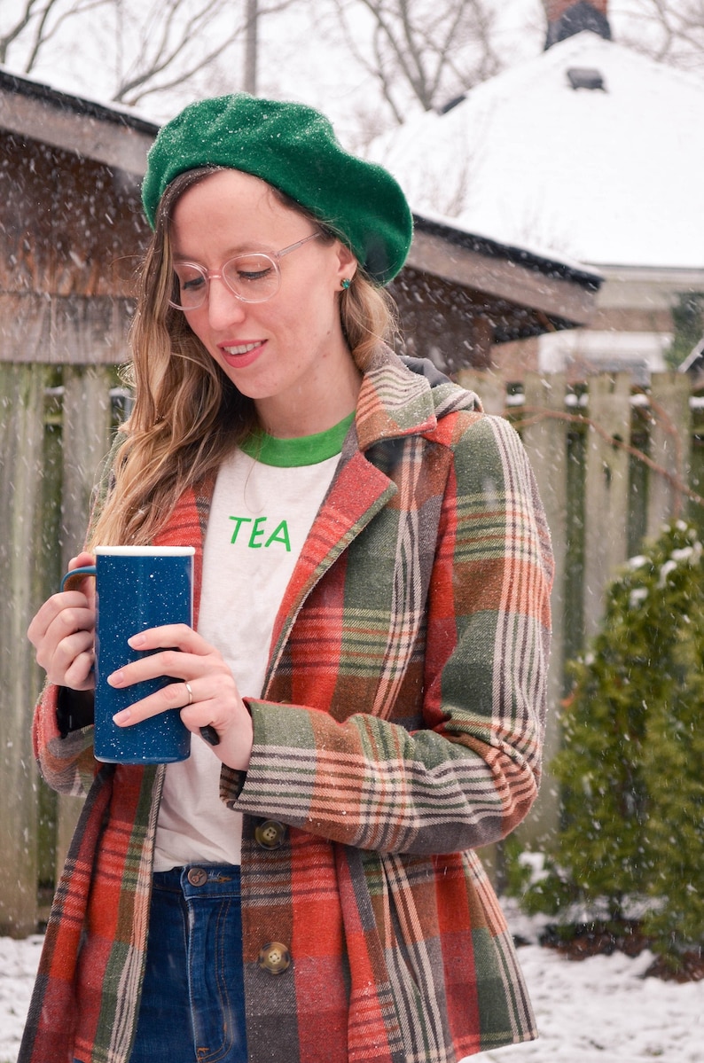 Light coloured, short sleeve t-shirt with bright ringer trim at neck and cuff screen printed to say Tea in matching colour print, modelled by woman in jeans, coat and beret in winter.