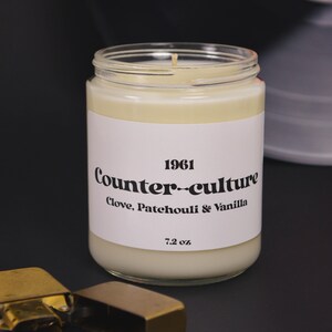 May include: A white candle in a glass jar with a white label that reads "1961 Counter-culture Clove, Patchouli & Vanilla 7.2 oz."