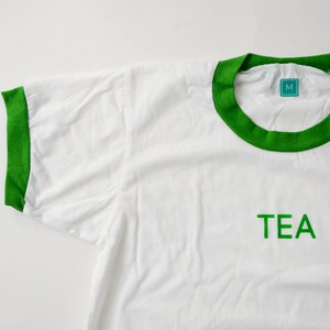 Light coloured, short sleeve t-shirt with bright ringer trim at neck and cuff screen printed to say Tea in matching colour print.