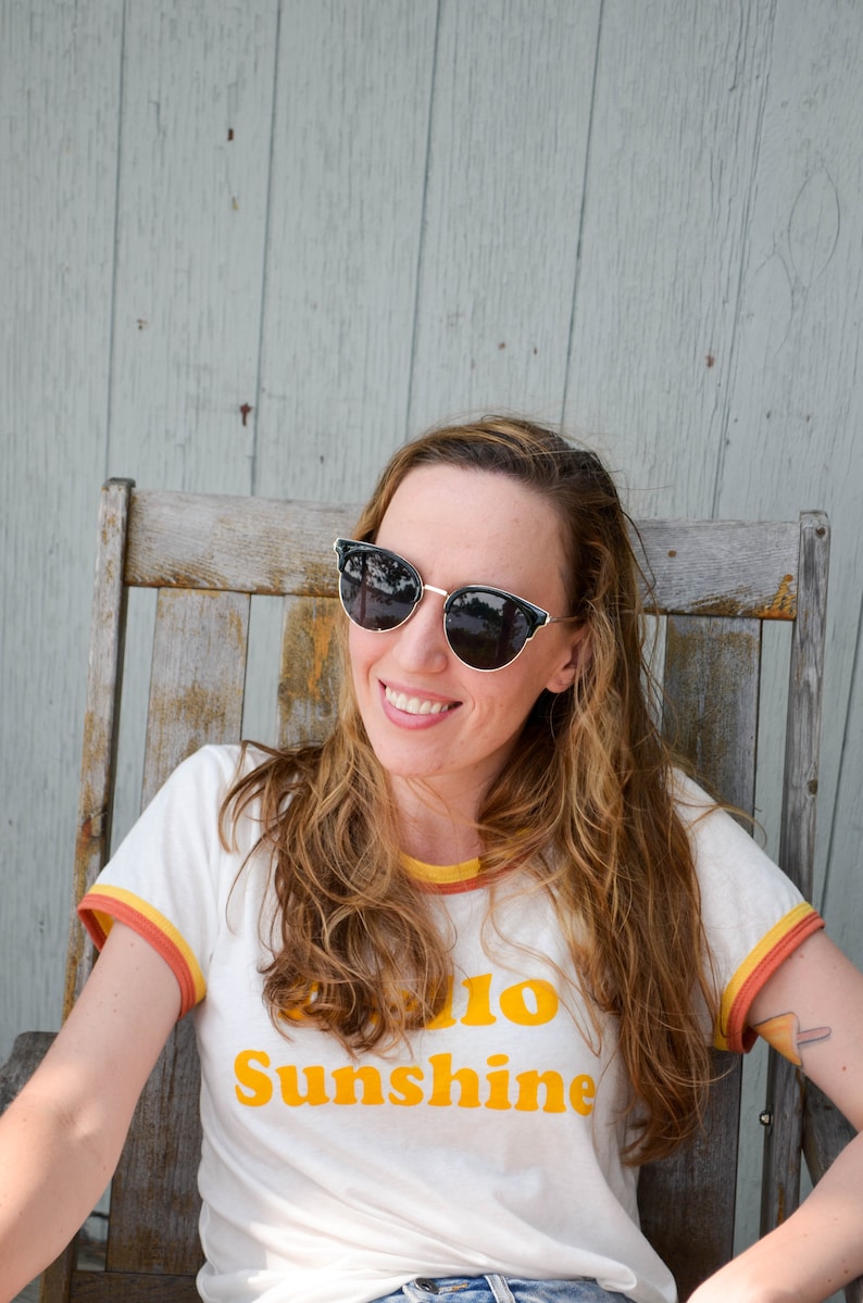 1970s retro style, loose fitting short sleeve t-shirt with colourful trim at neck and sleeves, screen printed to say Hello Sunshine in large font, modelled with denim shorts and sunglasses.