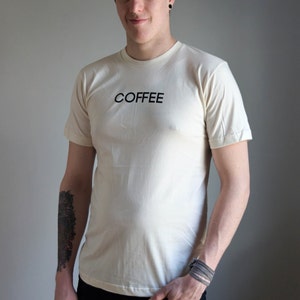 Light coloured, short sleeve t-shirt screen printed to say Coffee in dark font, modelled with dark pants.