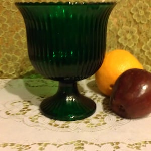 May include: A dark green glass goblet with vertical ribbed detailing. The goblet sits on a pedestal base. An orange and a red apple are next to the goblet on a white lace doily.