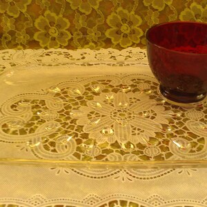 May include: A clear glass serving tray with a decorative pattern and a red glass cup on top. The tray has a raised edge and two handles on the sides.