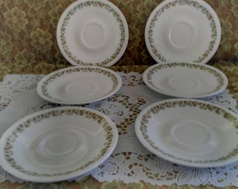 May include: Six white ceramic saucers with a green floral pattern around the edge. The saucers are arranged in a circle on a white lace tablecloth.