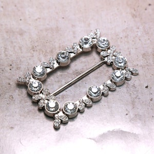 May include: A silver-toned rectangular buckle with a decorative design of clear rhinestones set in a floral pattern.