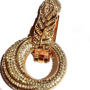 May include: A gold-toned clip-on earring with a feather-shaped top and a double-hoop design. The earring is encrusted with small, sparkling crystals, creating a glamorous and eye-catching accessory. The clip mechanism is visible.