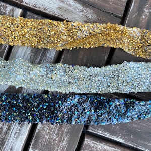 May include: Three strips of rhinestone trim in gold, clear, and iridescent black. The trim is made of small, faceted rhinestones.